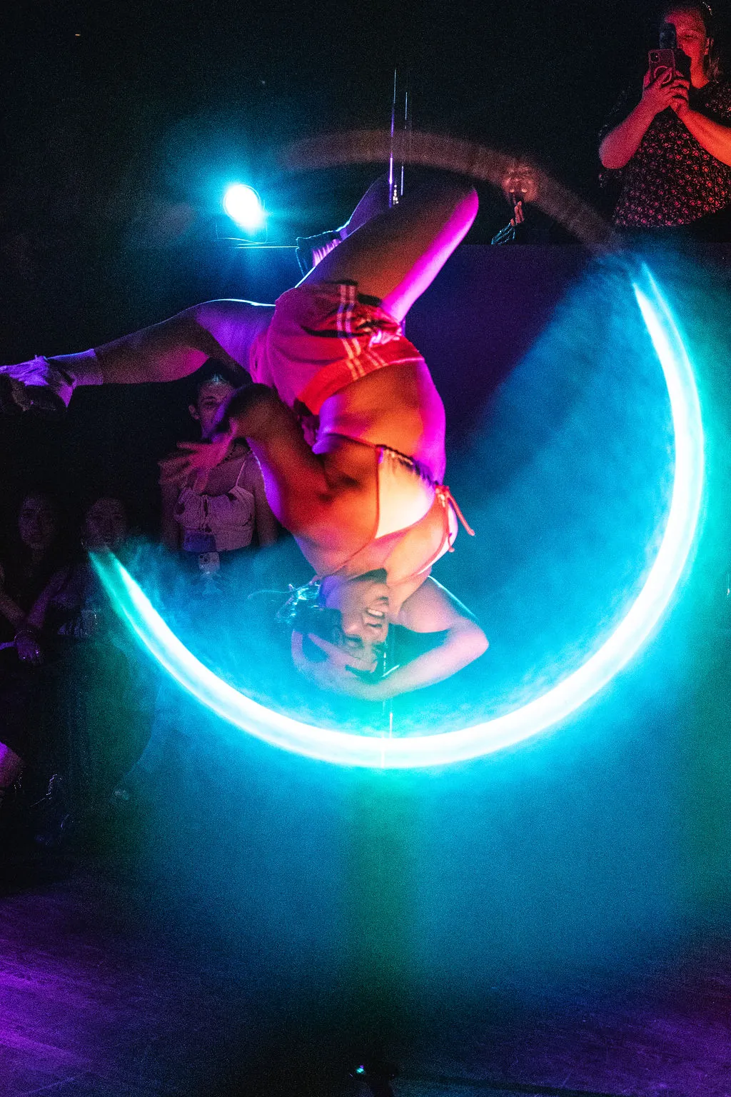 A performer inverted on the pole inside a ring of teal light, hair down, audience watching close at the edge of the floor