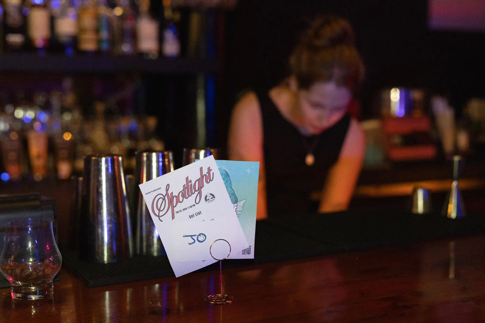 A set list card standing on the bar at Artemis, bartender working behind it