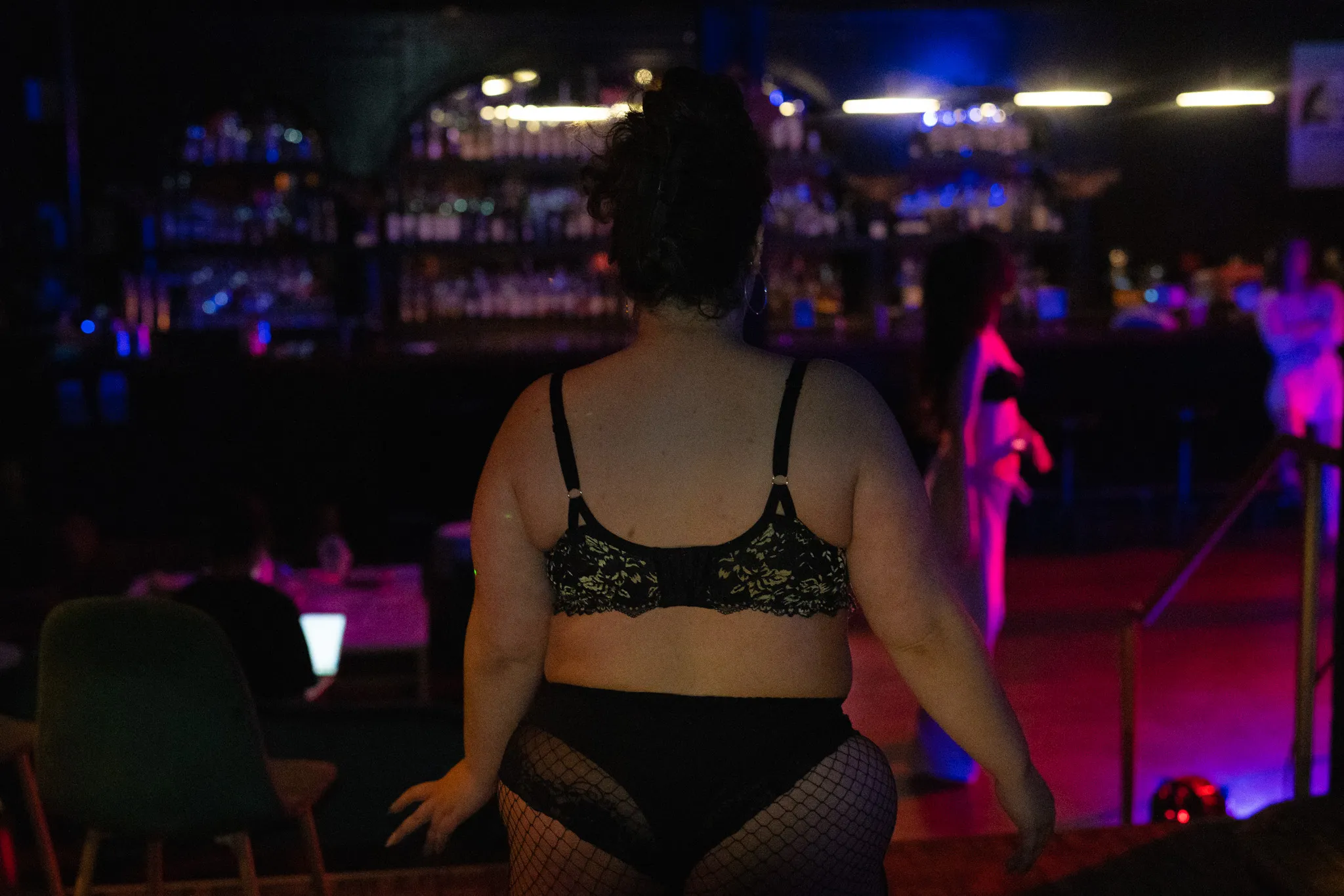 A performer waiting at the back of the room in the moments before going on, the bar glowing behind her